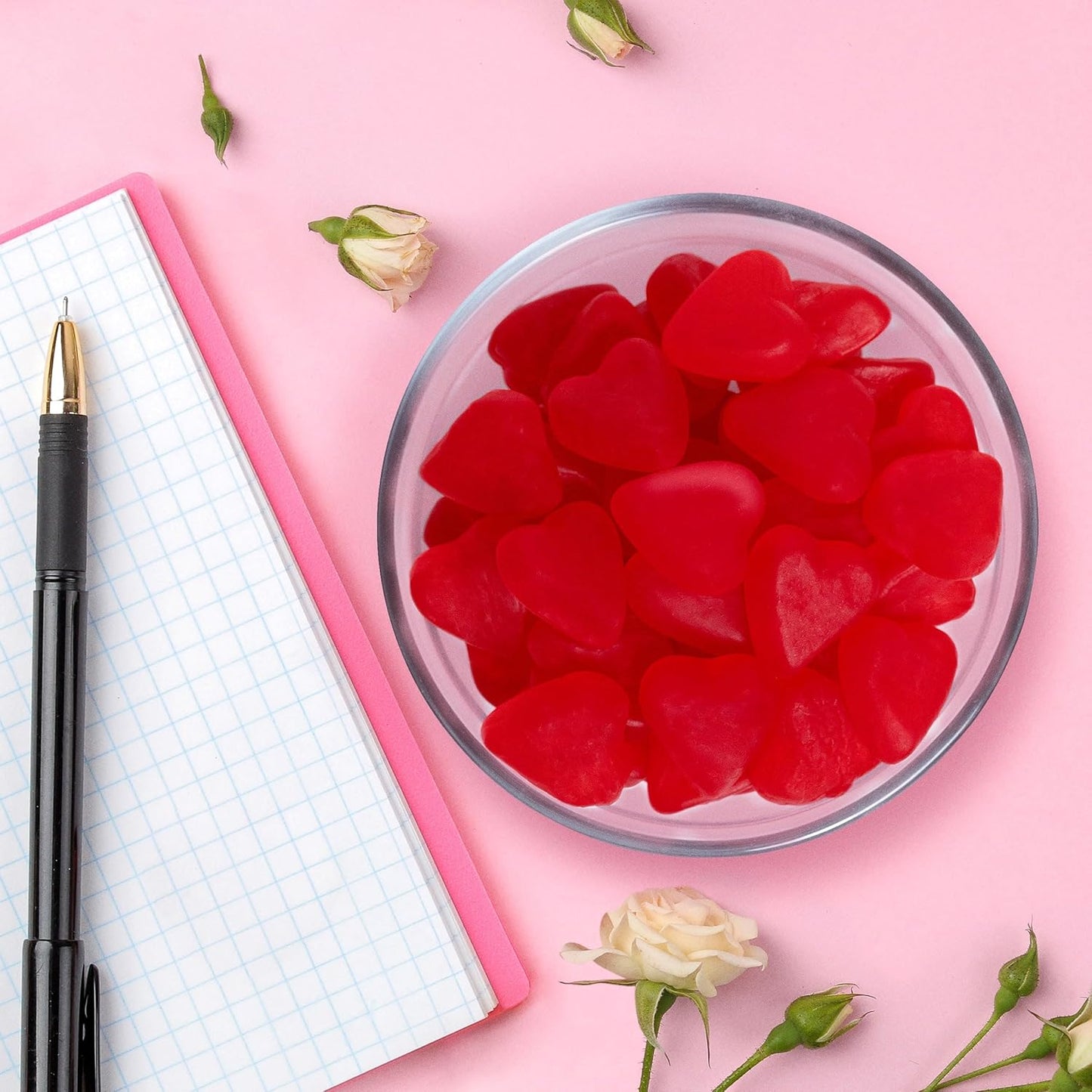 Red heart-shaped candies in a glass bowl on a pink surface with a pen and notebook.