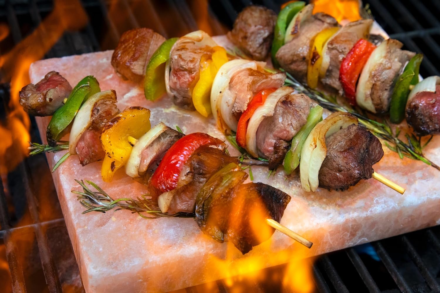 Skewers of meat and vegetables on a pink salt block over a grill with flames.