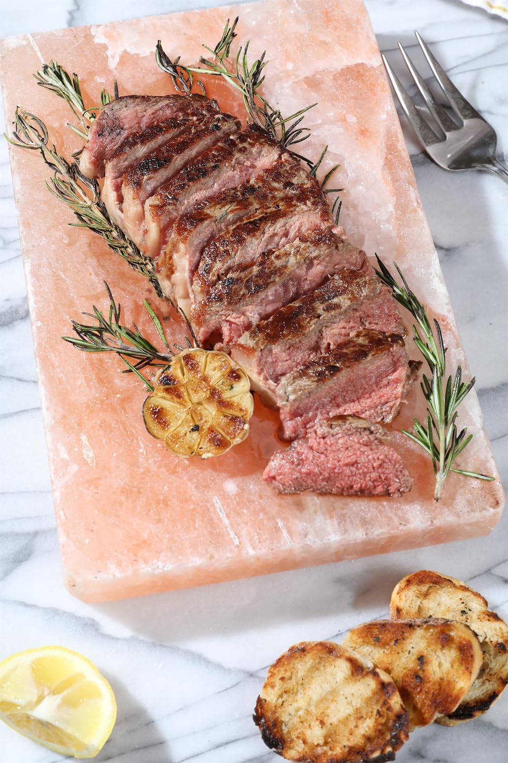 Sliced steak on a pink salt block with herbs and garlic, served on a marble surface.