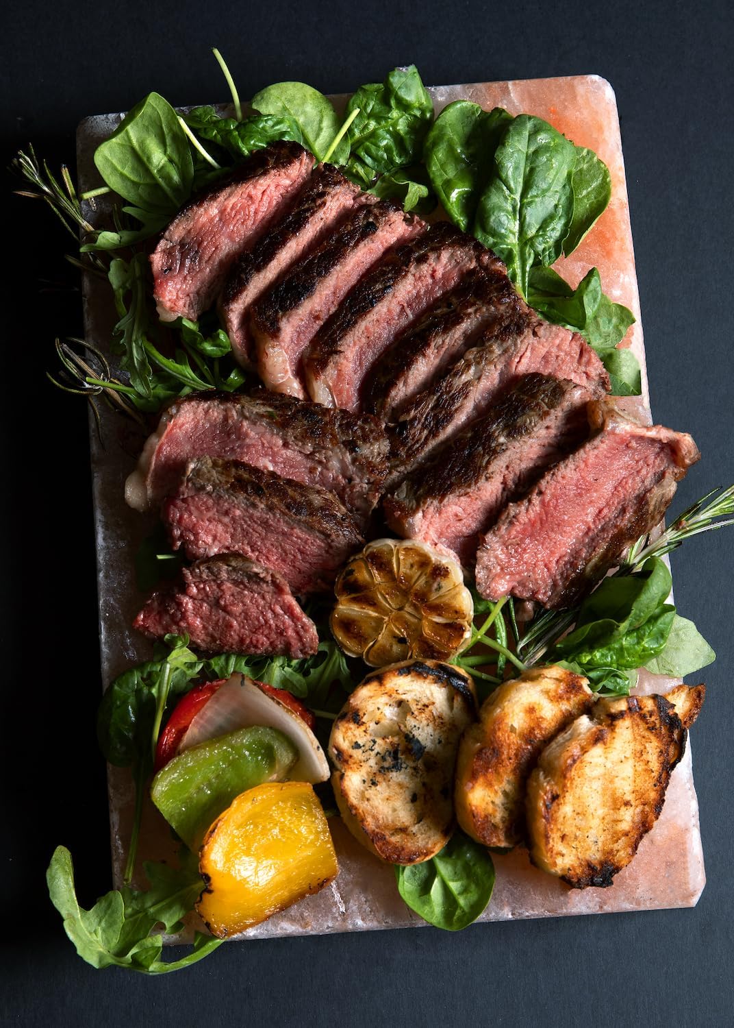 Sliced steak with vegetables on a pink salt block against a dark background