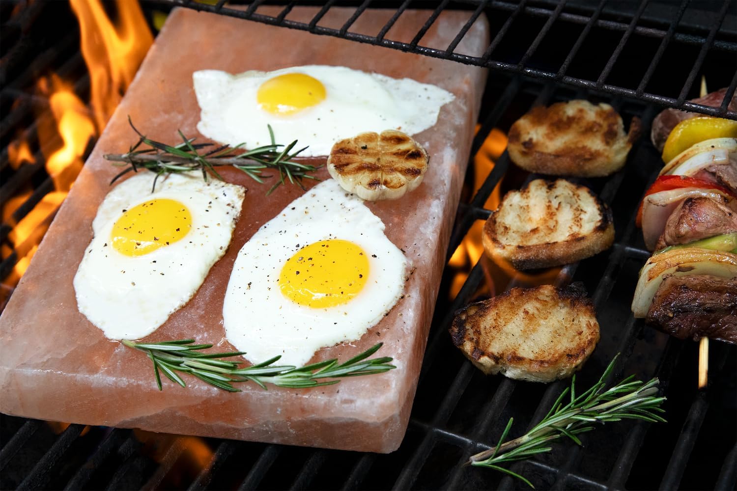 Fried eggs on a salt block with herbs, surrounded by grilled vegetables and meats on a barbecue.